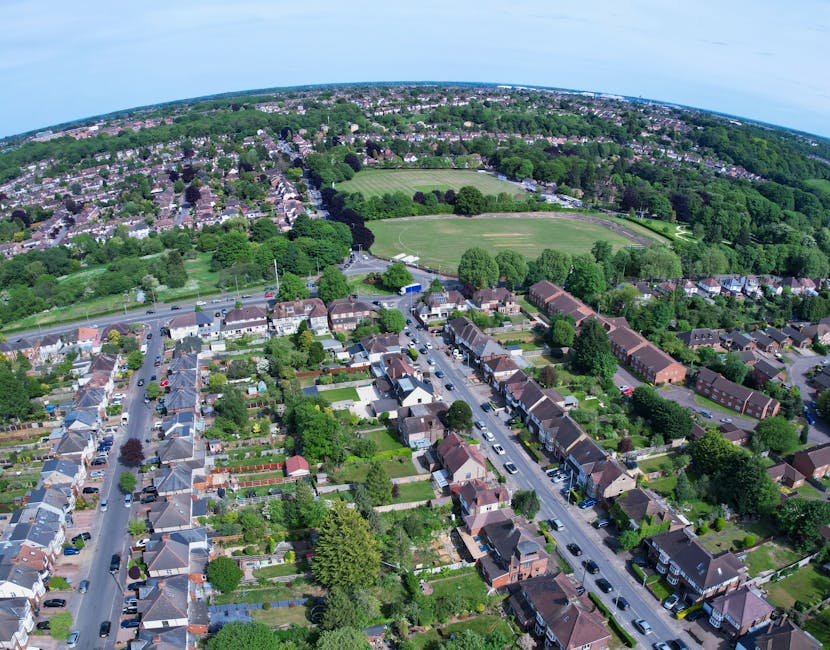An aerial view of a green, landscaped area featuring a mix of mature trees with lush foliage, some with white blossoms, and open grassy spaces. A narrow, winding dirt path weaves through the trees, pr