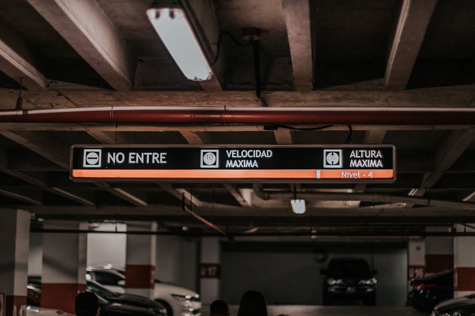A parking garage ceiling with a digital overhead sign displaying parking restrictions and speed limits in Spanish, including icons for no entry, maximum velocity of 10 km/h, and maximum height clearan